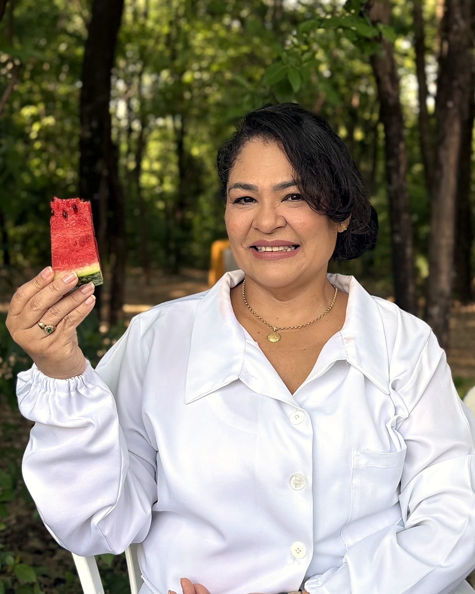 Retrato da nutricionista Márcia Glauciane, de jaleco branco, sorrindo ao ar livre com uma fatia de melancia na mão