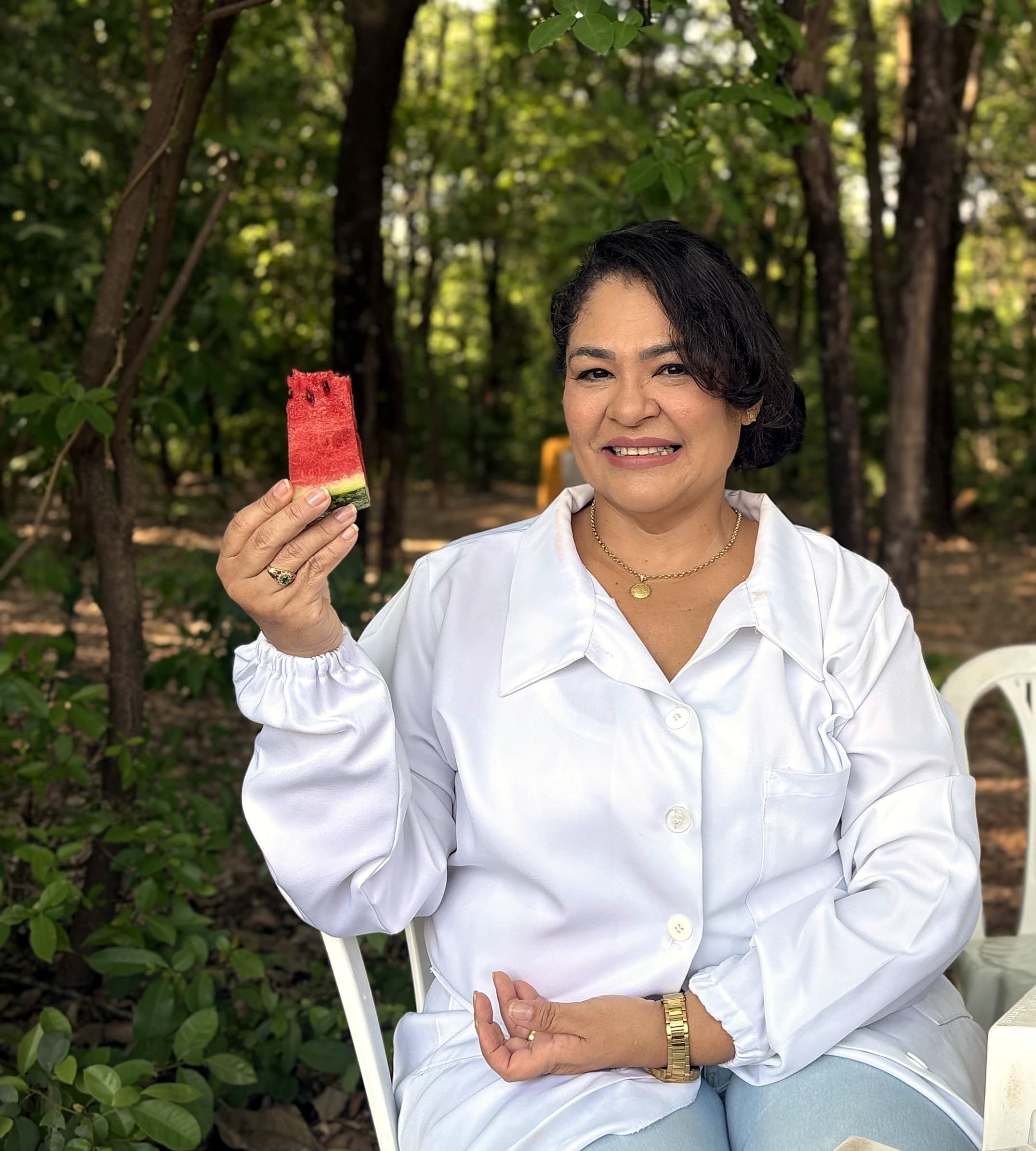 Márcia Glauciane, nutricionista, de jaleco branco, sorrindo e segurando uma fatia de melancia ao ar livre, entre árvores